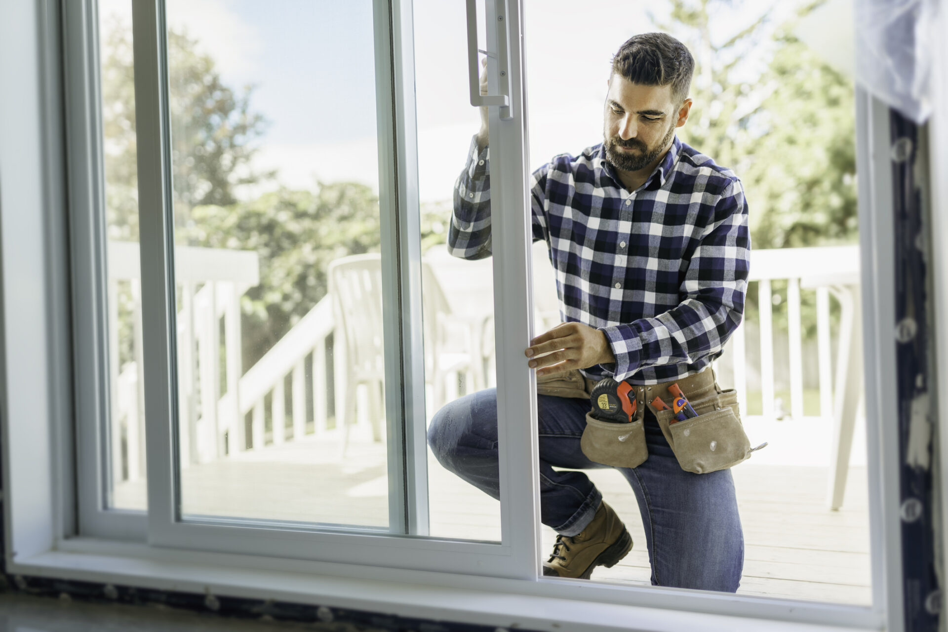 Man in plaid shirt with tool belt carefully installing energy-efficient sliding glass patio door in home, showcasing professional window and door replacement services.