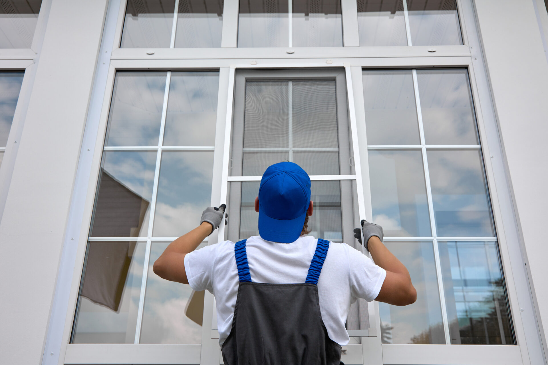 Technician in blue cap and gloves installing a window screen on a large white multi-pane window, demonstrating professional window replacement and energy-efficient home upgrades by a Buffalo-based union-made window company.